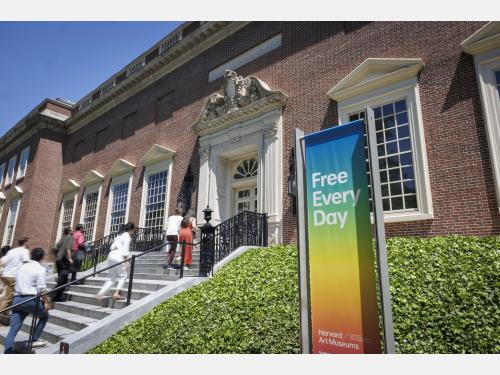 An exterior of a building with several people ascending a stone staircase into the building's entrance. Beside the staircase is a colorful sign that says "Free Every Day" in white text.