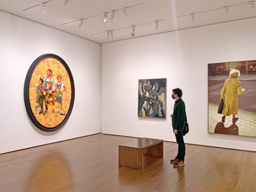 Person with face covering stands in a gallery looking at a large oval-shaped picture. 