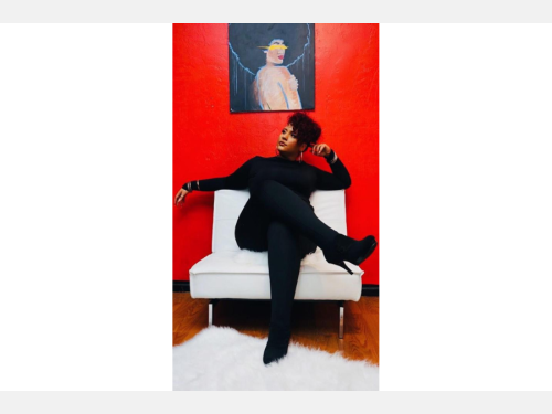 A Black woman sitting on a cushioned chair underneath a painting on a red wall.