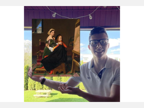A smiling young man, wearing glasses and a white shirt, stands near a picnic table, gesturing toward a floating painting. The painting shows an artist’s studio in which the artist, with a woman on his lap, looks over his shoulder at a painting in progress.