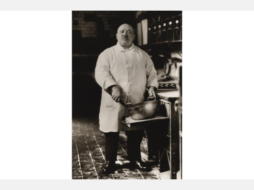 A portrait photograph of a bald white man standing in a kitchen mixing the contents of a metal bowl.