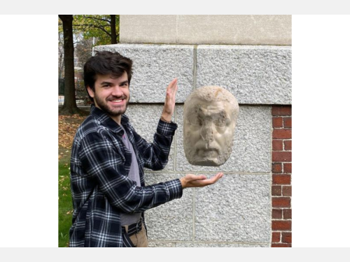 A smiling A smiling young man stands by a building, gesturing toward a marble head that appears to float.