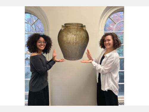 In this photomontage, two smiling young women stand indoors between tall windows, gesturing toward a mottled brown storage jar that appears to float between them.
