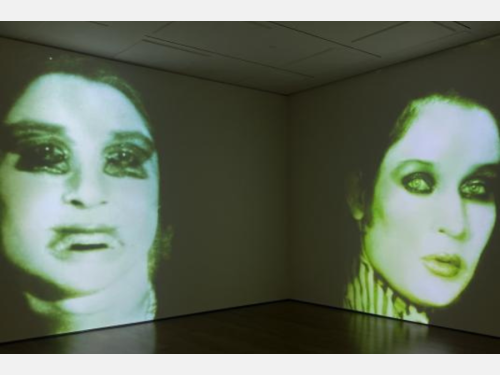 A gallery installation shot shows two close-up images of a woman’s face with neon green tones projected onto two walls.
