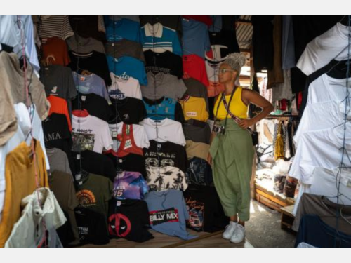 Photograph of a Black woman standing in front of T-shirts hanging on a wall. 