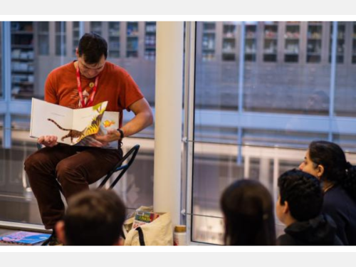 A man reads a picture book to a group of families in a gallery.