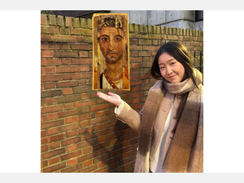 In this photomontage, a young woman, wearing a brown scarf and a beige winter jacket,  gestures toward a portrait that appears to float beside her. The portrait shows a lightly bearded young man and is painted on several slightly mismatched strips of material. 