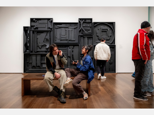 Two people happily chat on a bench with a large, black abstract sculpture behind them on a gallery wall.