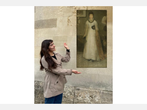 In this photomontage, a smiling young woman gestures toward a floating painting. It’s a full-length portrait of a ghostly young woman in a white dress, facing forward. The smiling woman wears a short trench coat and blue jeans and stands before a beige stone wall. 