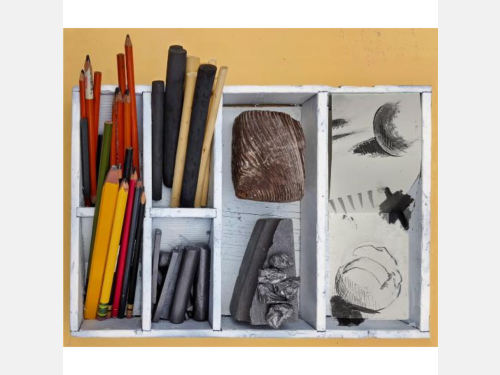 A white wooden box with six cubbyholes contains graphite and charcoal drawing implements, chunks of wood and graphite, and sample drawings.