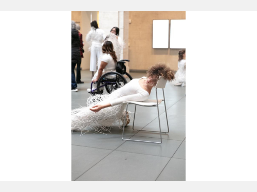 In an indoor courtyard, a performer dressed in all white and zip-tie wearable art is half-way slid off her chair. Another performer is posing in her wheelchair in the background.