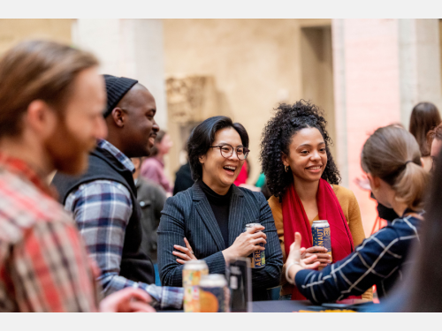 A group of people are gathered around a high table, holding drinks and smiling. The background is softly blurred.