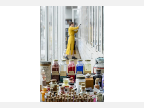 In the foreground of this photograph are several jars of pigments. In the background, a figure in yellow stands in a hallway.