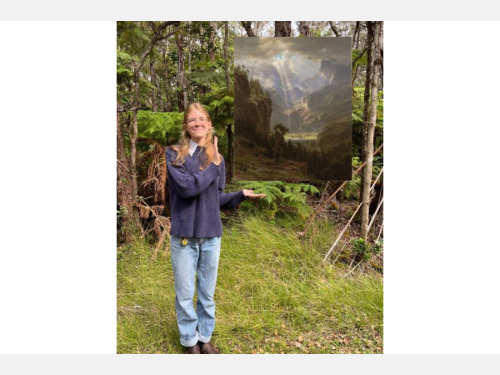 In this photomontage, a smiling young woman gestures toward a floating painting showing sunlight on a mountainous landscape. She stands in a wooded area wearing a blue sweater and blue jeans.