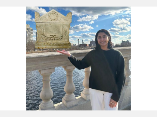 In this photomontage, a smiling young woman stands on a bridge gesturing toward a marble ash urn that resembles a small Roman temple with an arched roof, garlands, and an inscription on the facade. 