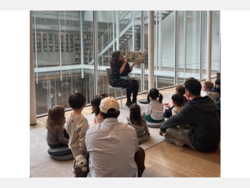 A woman reads to a group of children and caregivers in front of a glass window in a museum.