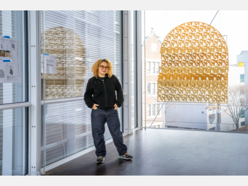 A woman stands in a gallery in front of a gold-colored metal sculpture with intricate, interlocking shapes; the sculpture is hanging from the ceiling, and a wall-sized window is behind it.