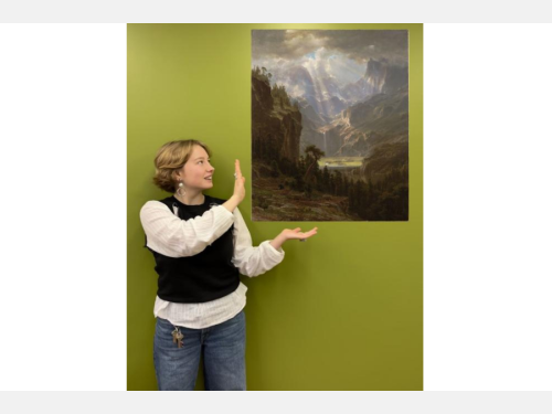 In this photomontage, a young woman gestures toward a painting that appears to float. It shows a sunlit valley surrounded by mountains and trees. The woman stands by a green wall wearing a white shirt, black vest, and blue jeans. 
