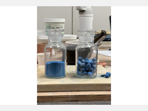 Two glass vials on a wooden surface are filled with blue pigment, one in powder form and one in small spheres.