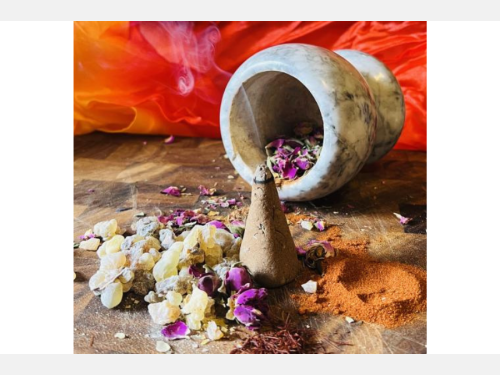Wisps of smoke rise from a cone-shaped incense, which is surrounded by frankincense resin, dried purple petals, and powdered spices laid out on a wooden surface. Behind the incense is a tipped marble mortar and a vivid orange-red fabric.