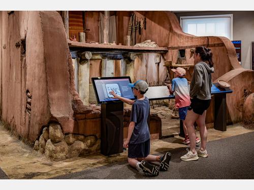 a woman and two children looking at a life-size replica of an ancient Mediterranean house