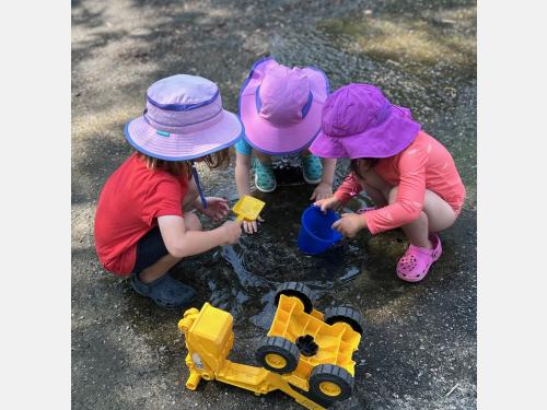 Photo of three children in summer hats circled around a pool of water with a cup and shovel in hand.